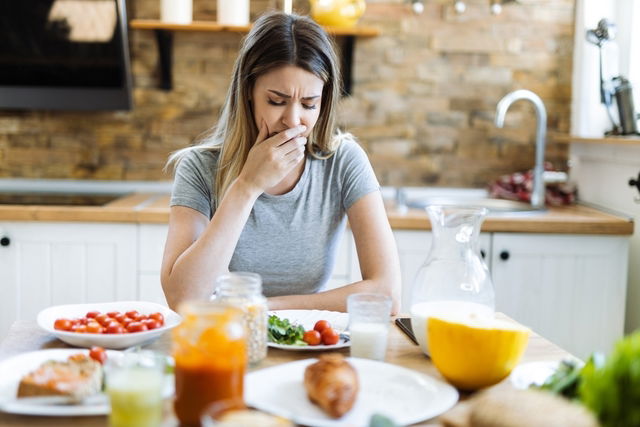 Mujer joven sentada en la mesa con diferentes alimentos, sintiendo dolor y náuseas al comer