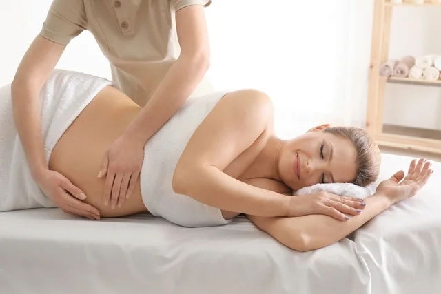 Mujer embarazada recostada de costado en una cama mientras le hacen masajes en la barriga