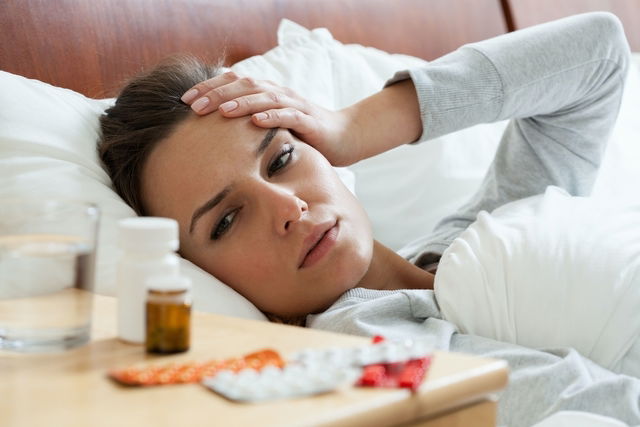 Mujer recostada en una cama con dolor de cabeza y medicamentos apoyados en la mesa