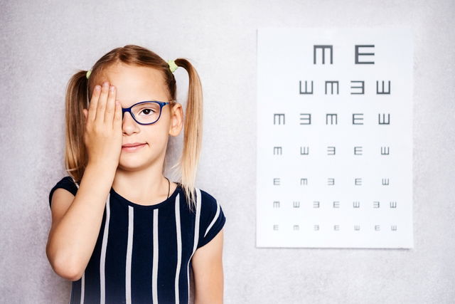 Niña pequeña que usa anteojos que se hace la prueba de vista antes de la escuela con una gráfica de 